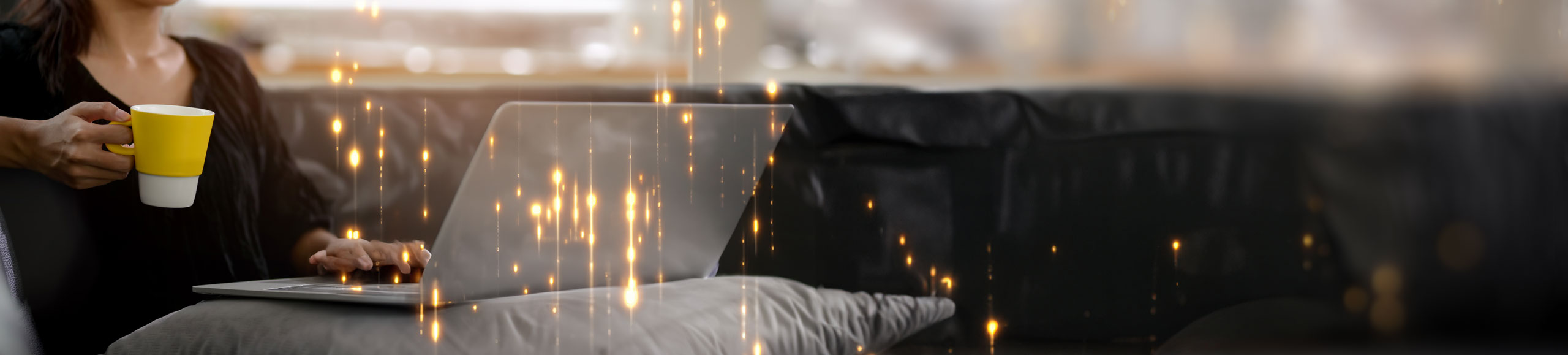 Woman holding a yellow coffee mug while using a laptop on a black sofa.