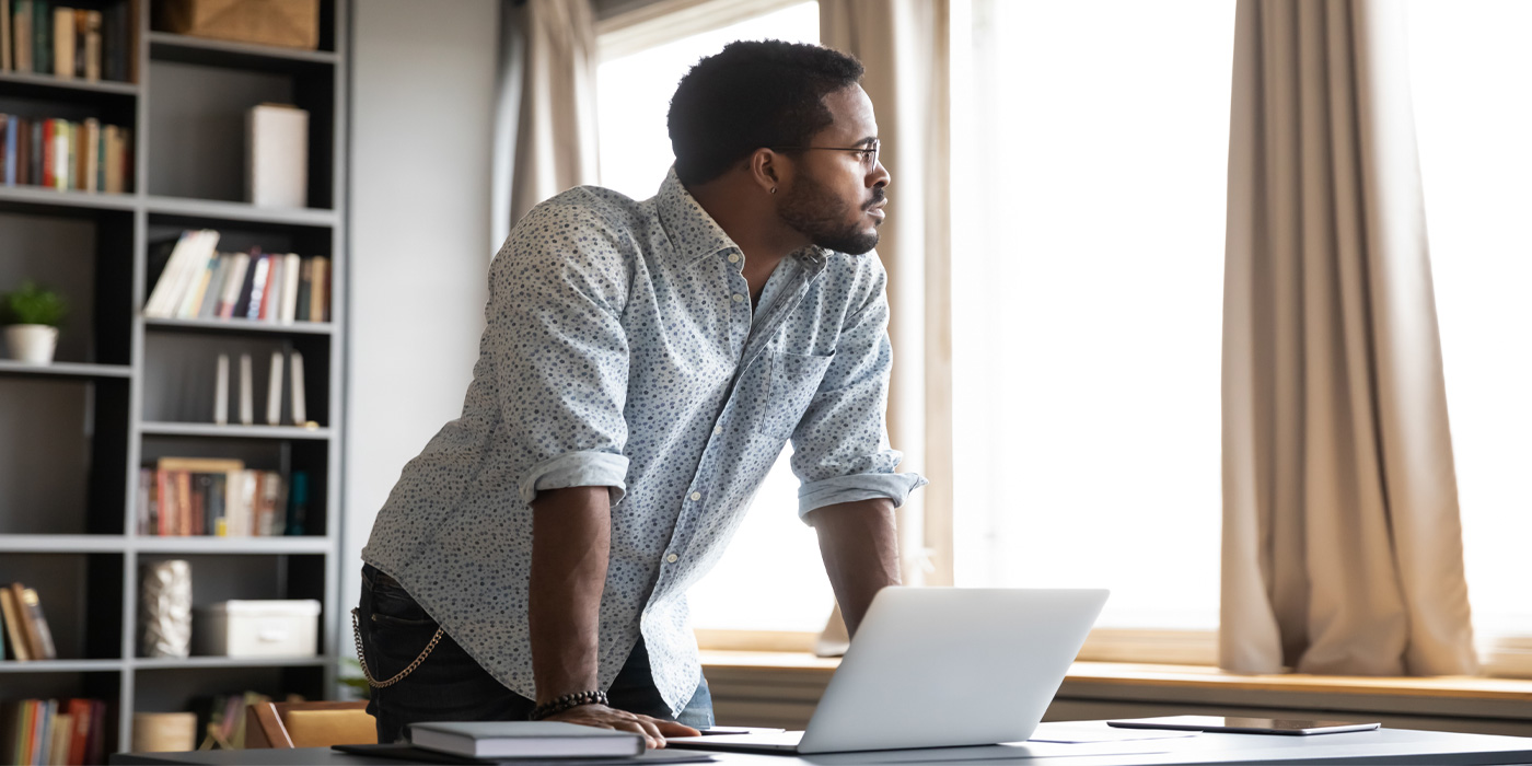 Man in casual shirt standing by a window with a laptop, looking thoughtful.