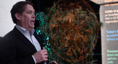 Man speaking at a conference with informative graphics in the background.