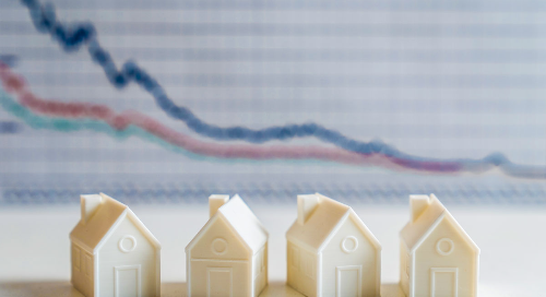 Miniature houses with declining graph in the background indicating real estate market trends.