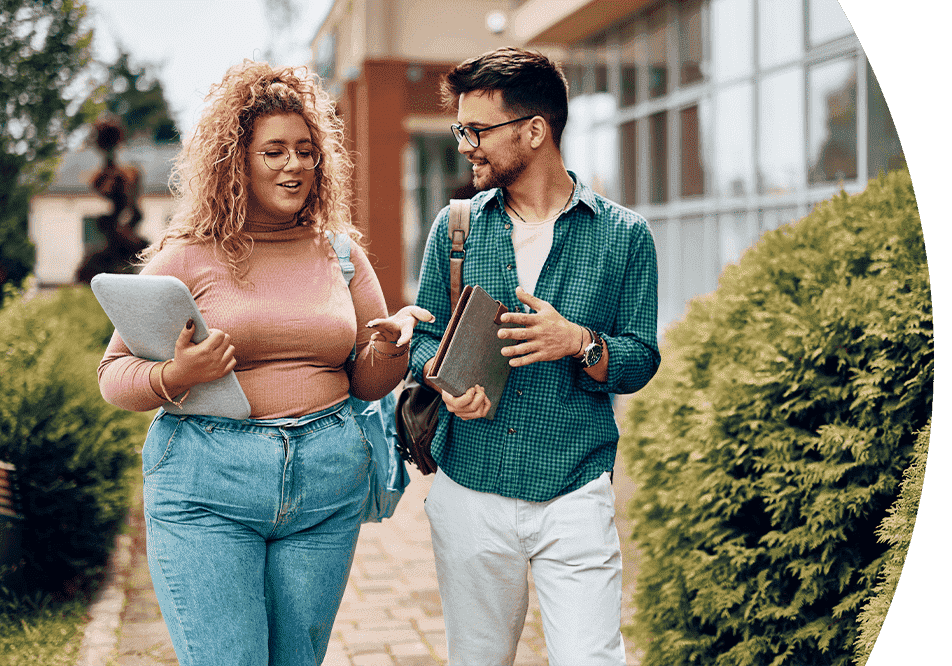 HigherEducation_MaximizeEnrollment Two young adults walking and talking with books and laptops on a college campus.