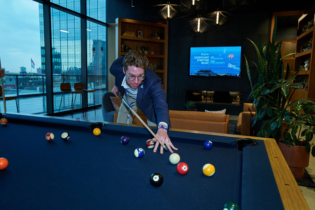 L1081550 Man playing pool in modern office with city view and TV in background.