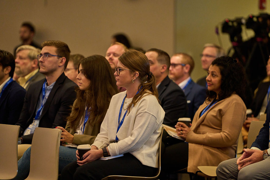 _P1_3173 Audience attentively listening at a business conference with lanyards visible.
