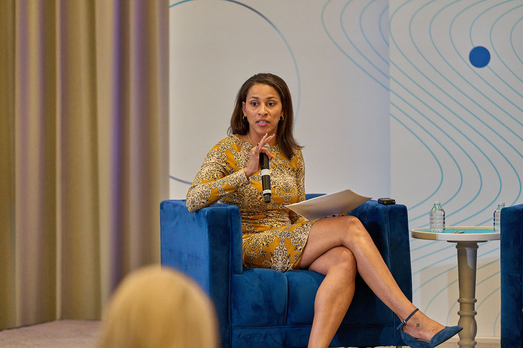 _P1_3722 Woman in yellow dress speaking at a conference, sitting on a blue chair with a microphone in hand.