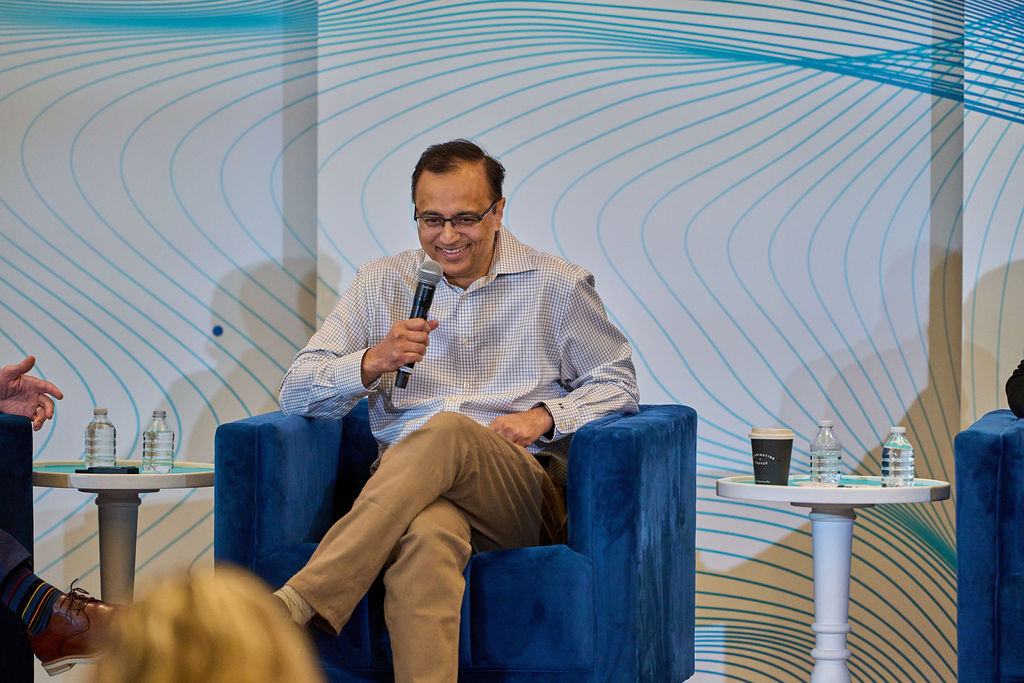 _P1_3923 Man speaking into a microphone while seated on a blue chair, with patterned backdrop.