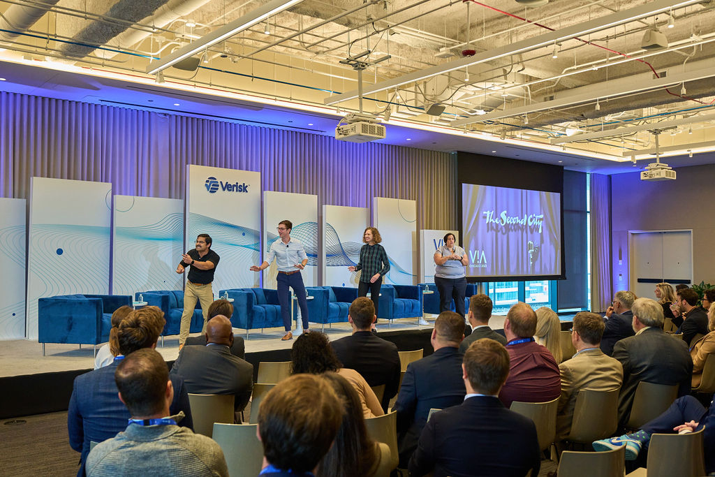 _P2_8089 Four presenters on stage at a Verisk conference engaging with a seated audience.
