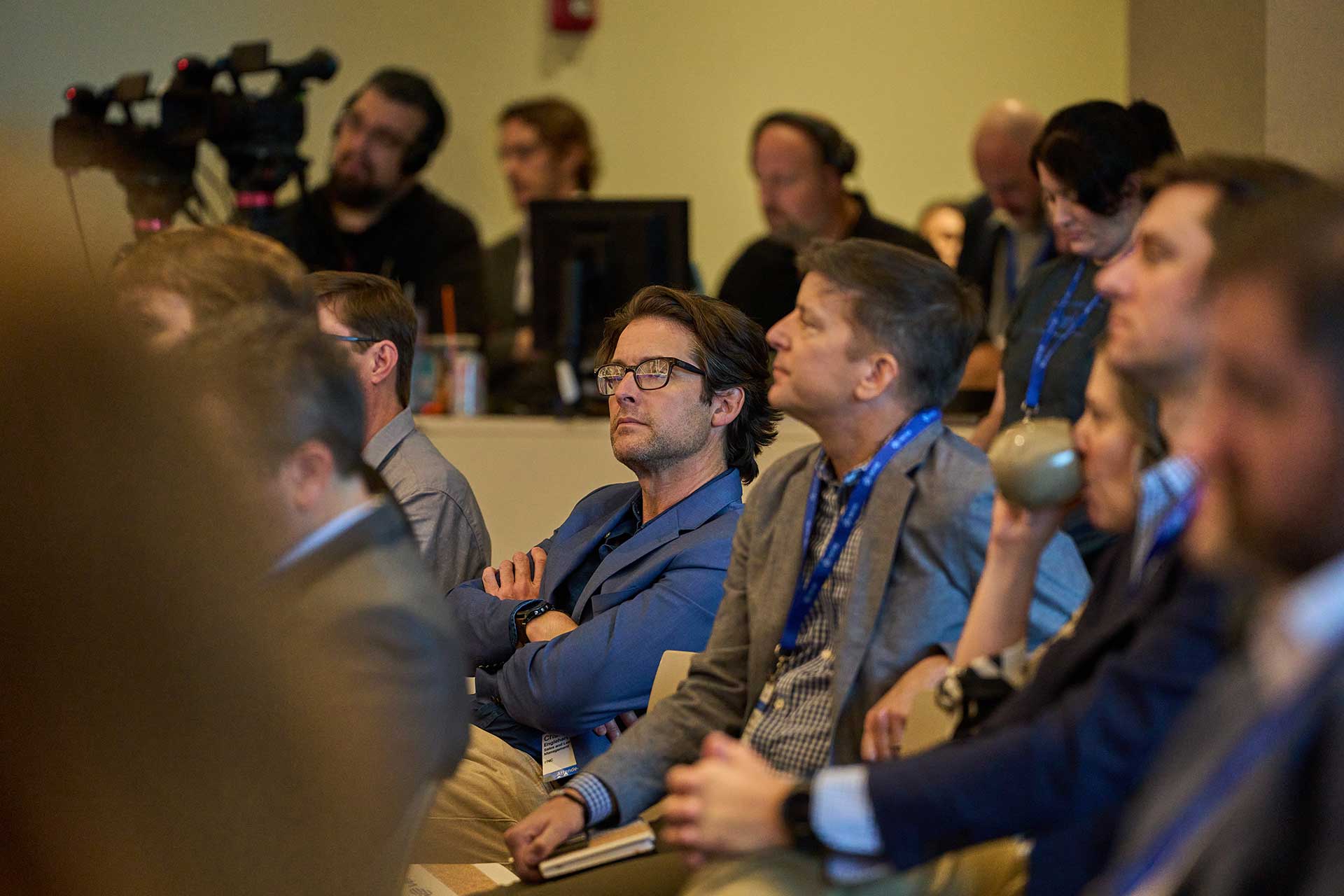 Group of conference attendees listening attentively with media crew in the background.